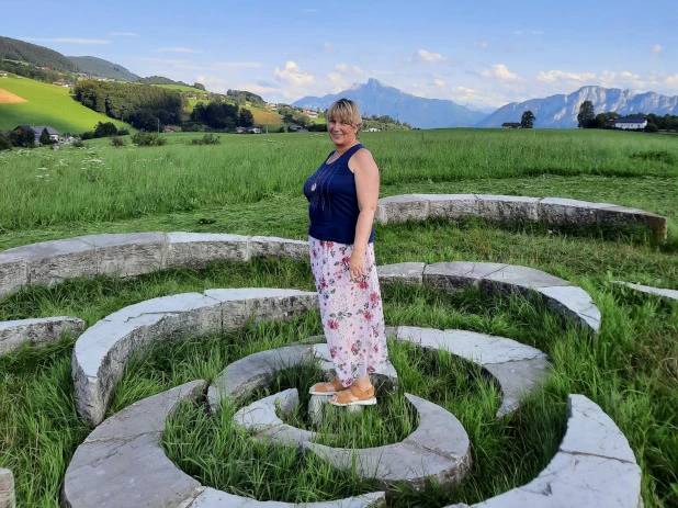 Frau in Blumenrock steht auf einem Steinkreis vor einer weiten Grünlandschaft und Bergkulisse